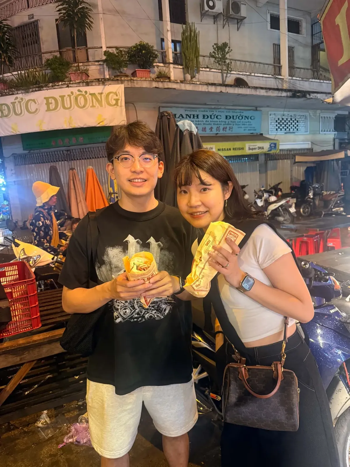 Two travellers holding fresh bánh mì on a busy street at night during a Saigon food tour